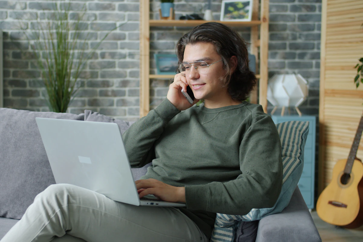 A homeowner on the phone with a laptop, relaxing on the couch in a brick-walled city apartment